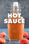 Clear glass bottle of hot pepper hot sauce filled with vibrant orange liquid, surrounded by red peppers on a blue surface. The text on the image reads, “Honey Roasted Hot Pepper HOT SAUCE.” Website: chilipeppermadness.com.