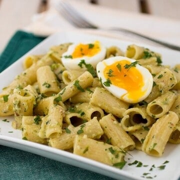 A plate of rigatoni pesto pasta recipe topped with chopped herbs, sliced almonds, and a soft-boiled egg cut in half, served on a white square dish atop a green napkin.