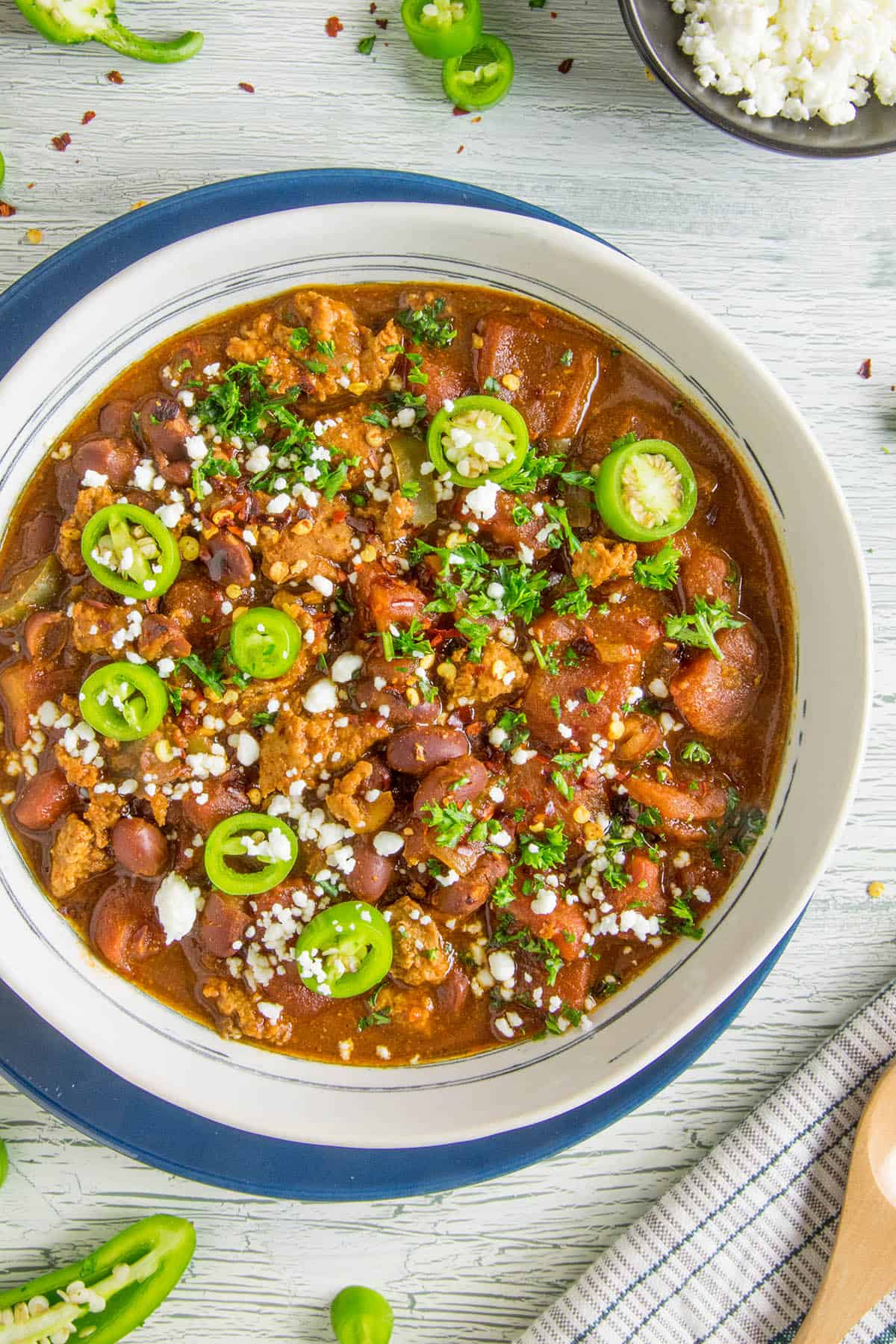 A finished bowl of chili, with sliced jalapenos, chopped herbs and crumbly white cheese on top