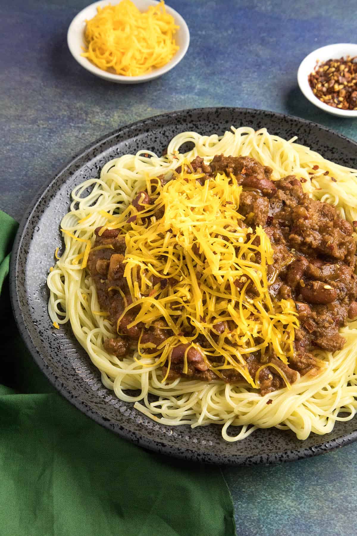 Cincinnati Chili Recipe Cincinnati Chili on a plate of noodles with shredded cheddar cheese over the top, ready to eat