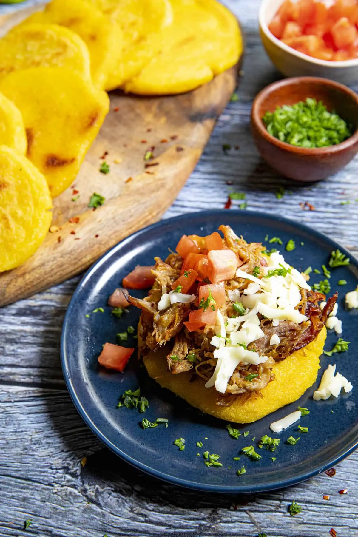 An arepa topped with pulled pork, shredded cheese and diced tomato with an extra stack of arepas