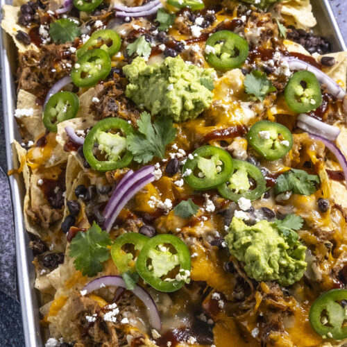 Serving up pulled pork nachos on a large tray, loaded with toppings, including guacamole, sliced jalapenos, sliced red onion, and bbq sauce
