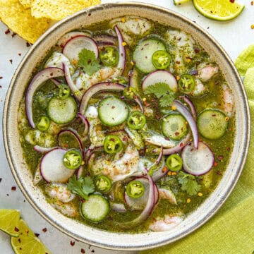 A bowl of aguachile features shrimp, cucumber slices, red onions, and cilantro in a vibrant green sauce. Lime slices and tortilla chips accompany this delicious aguachile recipe on a light surface with a green cloth.