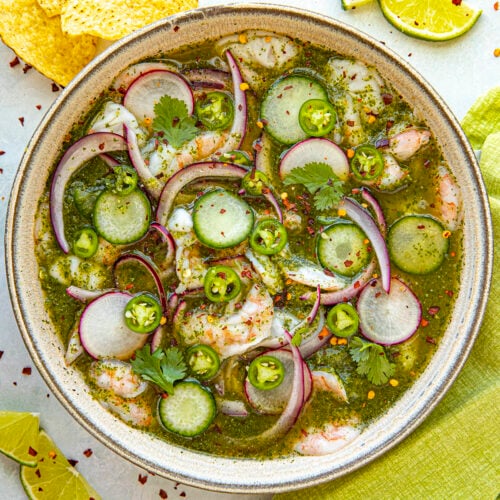A bowl of aguachile features shrimp, cucumber slices, red onions, and cilantro in a vibrant green sauce. Lime slices and tortilla chips accompany this delicious aguachile recipe on a light surface with a green cloth.
