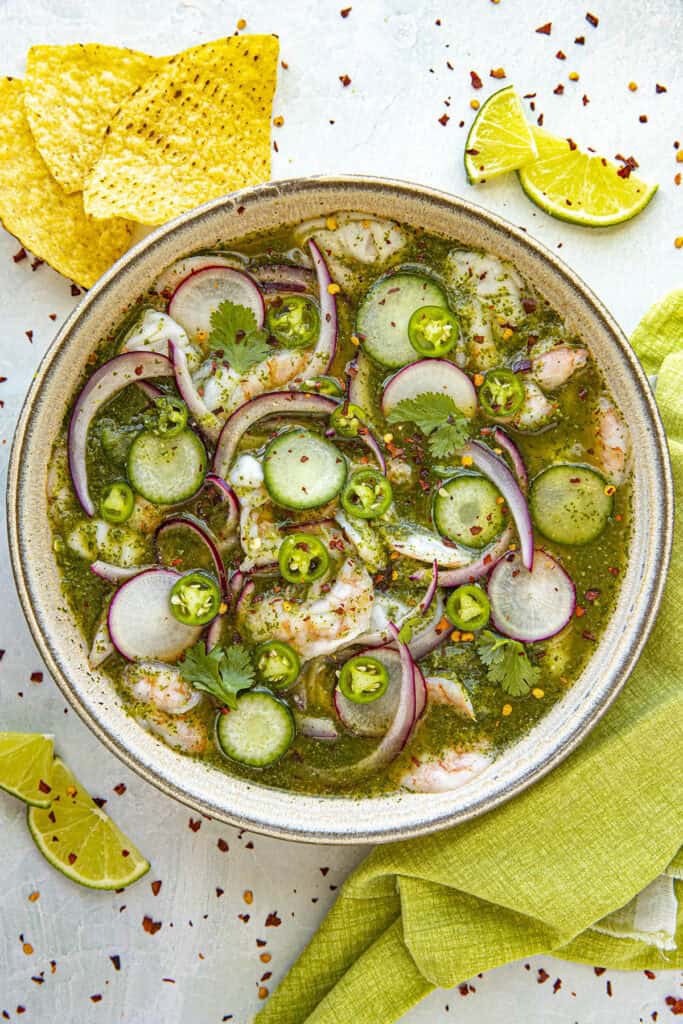 A bowl of aguachile features shrimp, cucumber slices, red onions, and cilantro in a vibrant green sauce. Lime slices and tortilla chips accompany this delicious aguachile recipe on a light surface with a green cloth.