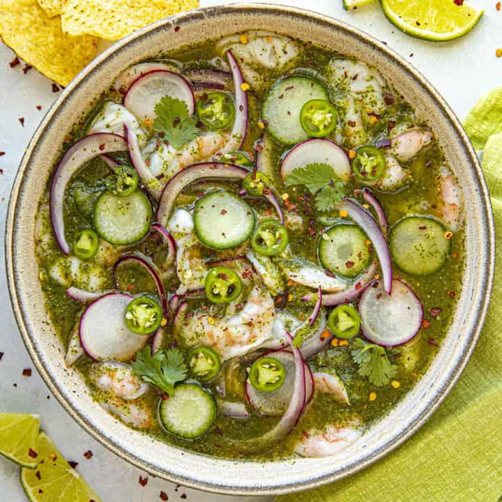 A bowl of aguachile features shrimp, cucumber slices, red onions, and cilantro in a vibrant green sauce. Lime slices and tortilla chips accompany this delicious aguachile recipe on a light surface with a green cloth.
