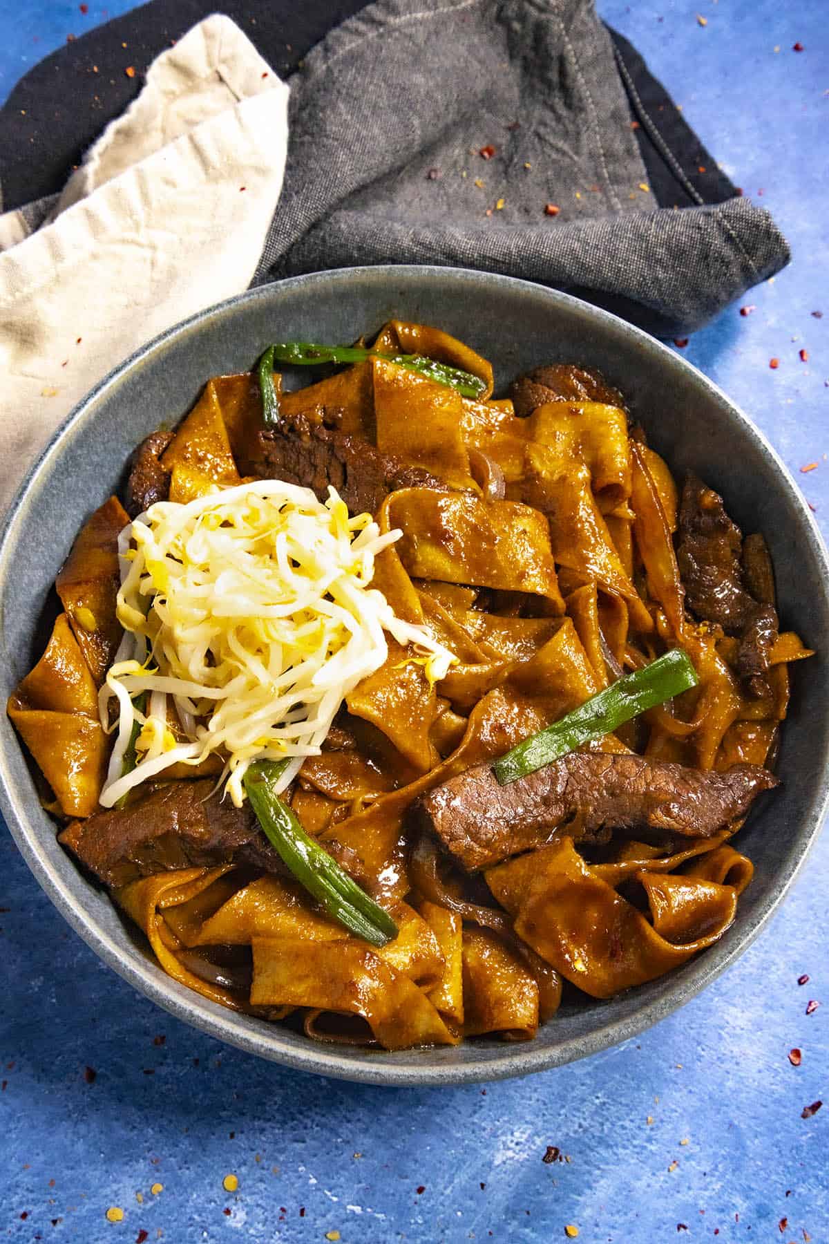 A bowl of beef chow fun, featuring wide rice noodles stir-fried with tender beef, is garnished with bean sprouts and green onions. Served in a blue bowl on a matching surface, the dish captures a rustic charm against the backdrop of dark and light cloth.
