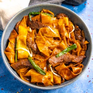 A bowl of wide, flat noodles mixed with slices of beef, green onions, and bean sprouts offers a delicious beef chow fun experience. The dish is coated in a rich, dark sauce.