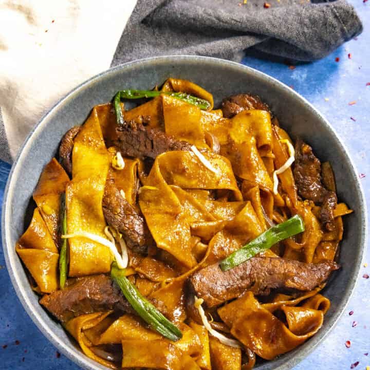 A bowl of beef chow fun features wide, flat noodles with tender beef slices, garnished with green onions. The richly sauced dish boasts a glossy texture. Beside the bowl on a blue surface lies a folded dark cloth napkin accented with a lighter section.
