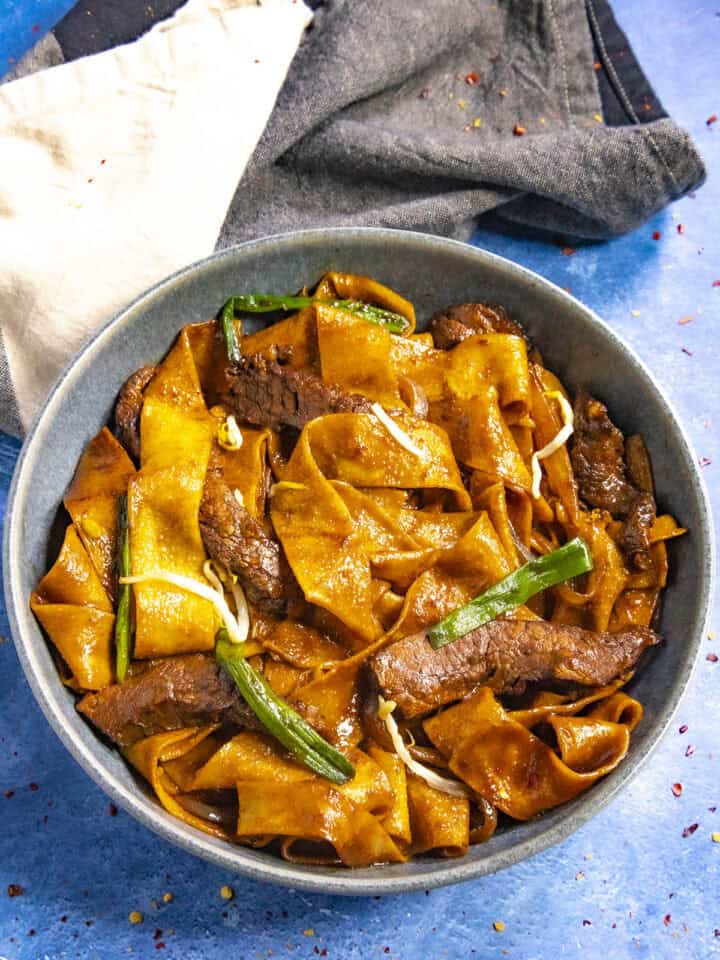 A bowl of beef chow fun features wide, flat noodles with tender beef slices, garnished with green onions. The richly sauced dish boasts a glossy texture. Beside the bowl on a blue surface lies a folded dark cloth napkin accented with a lighter section.