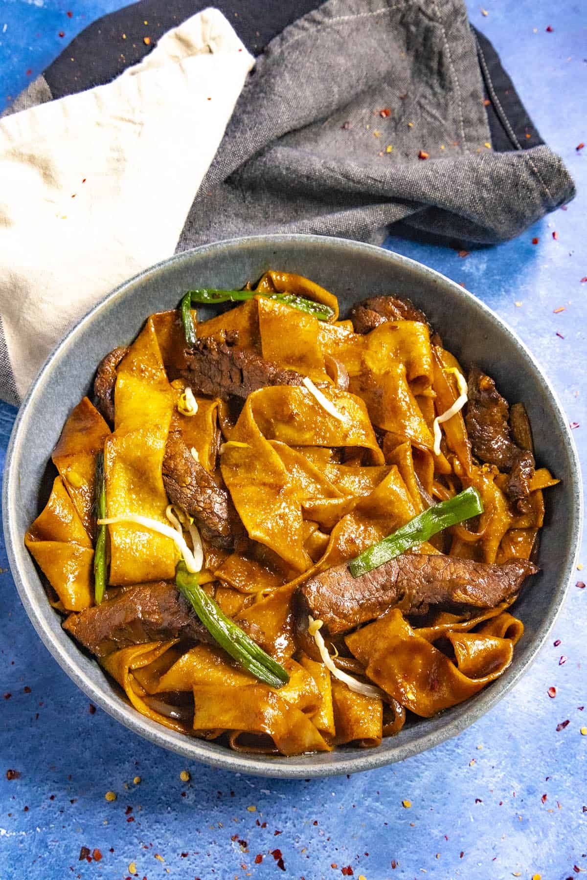 A bowl of beef chow fun features wide, flat noodles with tender beef slices, garnished with green onions. The richly sauced dish boasts a glossy texture. Beside the bowl on a blue surface lies a folded dark cloth napkin accented with a lighter section.