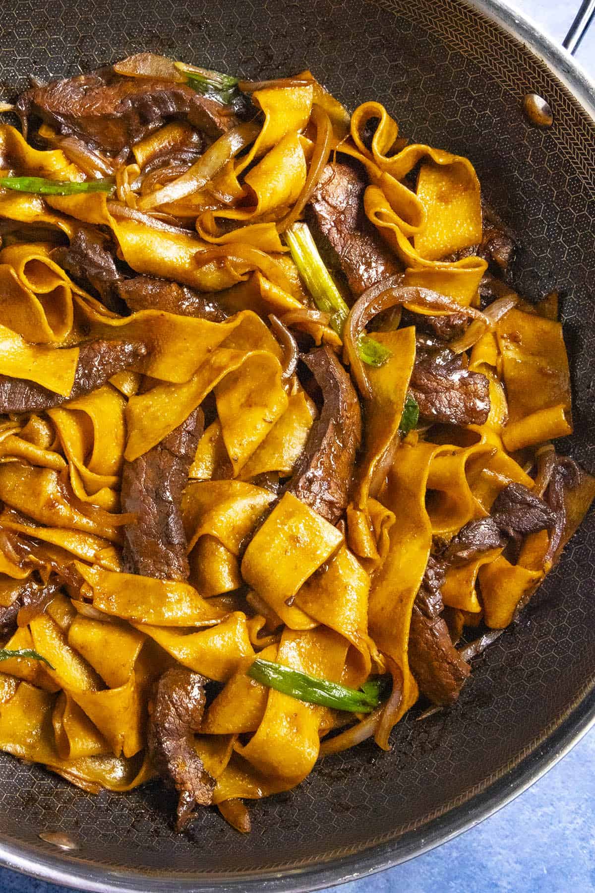 A pan full of wide, shiny chow fun noodles mixed with slices of meat and garnished with green onions. This Beef Chow Fun Recipe is saucy, boasting a rich brown color.