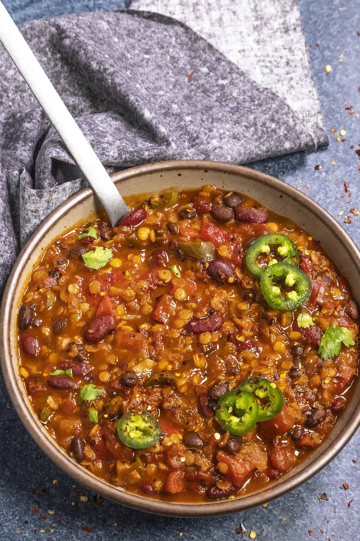 A bowl of lentil chili filled with beans and tomatoes is garnished with slices of jalapeño and cilantro. A spoon rests in the bowl, and a gray napkin is placed beside it on a textured blue surface.