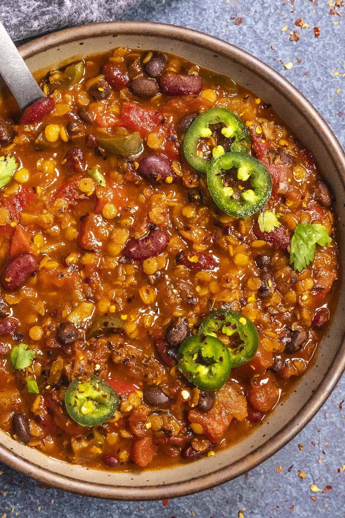 A bowl of lentil chili filled with lentils, beans and tomatoes, garnished with sliced jalapeños and cilantro. A spoon rests inside the bowl, which is placed on a textured surface with a cloth nearby.