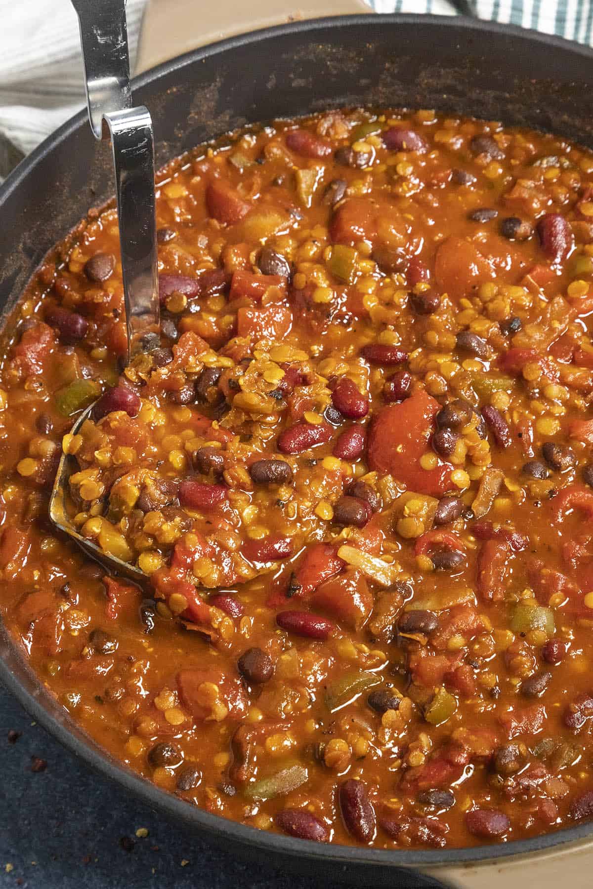 A close-up of a pot filled with hearty lentil chili. The chili consists of kidney beans, black beans, tomatoes, and lentils, mixed in a rich, red sauce. A metal ladle rests in the pot, ready to serve.