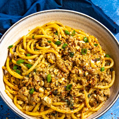 Dan Dan Noodles in a bowl, ready to serve, garnished with roasted peanuts, sesame seeds, chili flakes, and sliced green onion