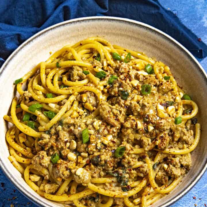 Dan Dan Noodles in a bowl, ready to serve, garnished with roasted peanuts, sesame seeds, chili flakes, and sliced green onion