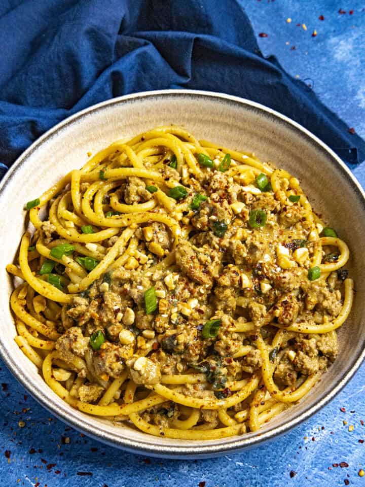 Dan Dan Noodles in a bowl, ready to serve, garnished with roasted peanuts, sesame seeds, chili flakes, and sliced green onion