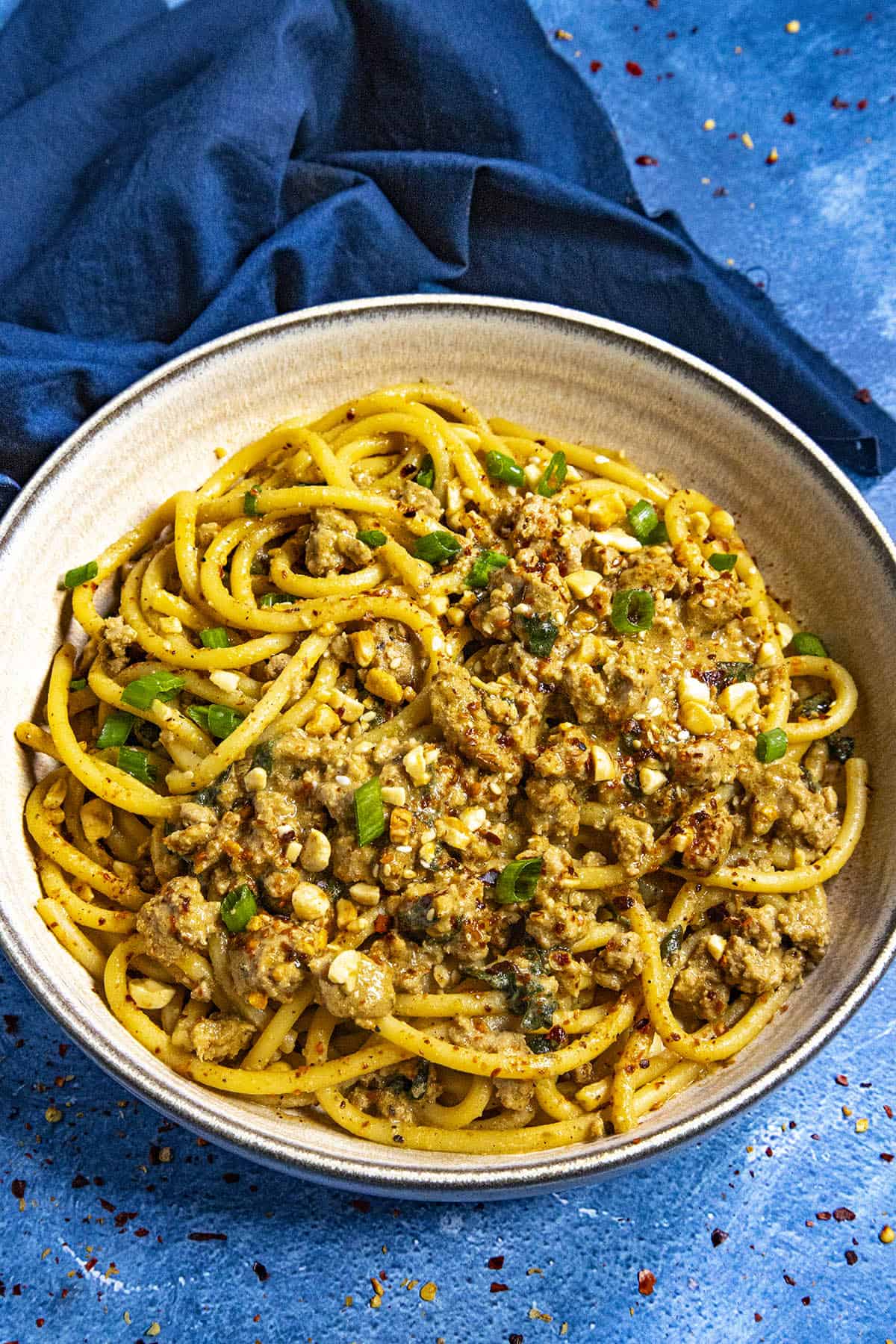 Dan Dan Noodles in a bowl, ready to serve, garnished with roasted peanuts, sesame seeds, chili flakes, and sliced green onion