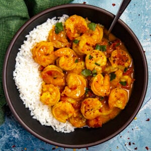 A bowl of white rice topped with shrimp in a yellow, spiced sauce with chopped green herbs and red pepper flakes, served on a blue surface with a green napkin nearby.