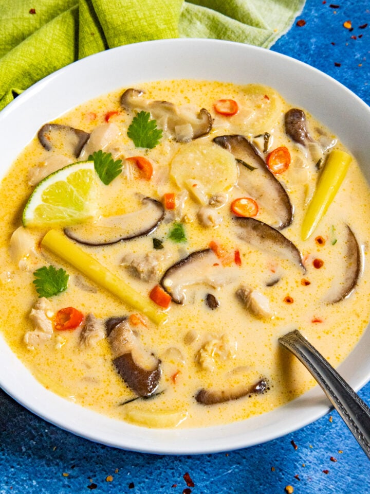 A bowl of creamy Thai coconut chicken soup, or tom kha gai, with sliced mushrooms, chili peppers, lemongrass, a lime wedge, and cilantro sits on a blue surface. A spoon rests in the bowl while a green cloth adds contrast in the background.