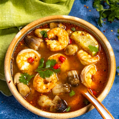 A large bowl of spicy Tom Yum Soup loaded up with shrimp and mushrooms, garnished with spicy red chilies, cilantro, and lime