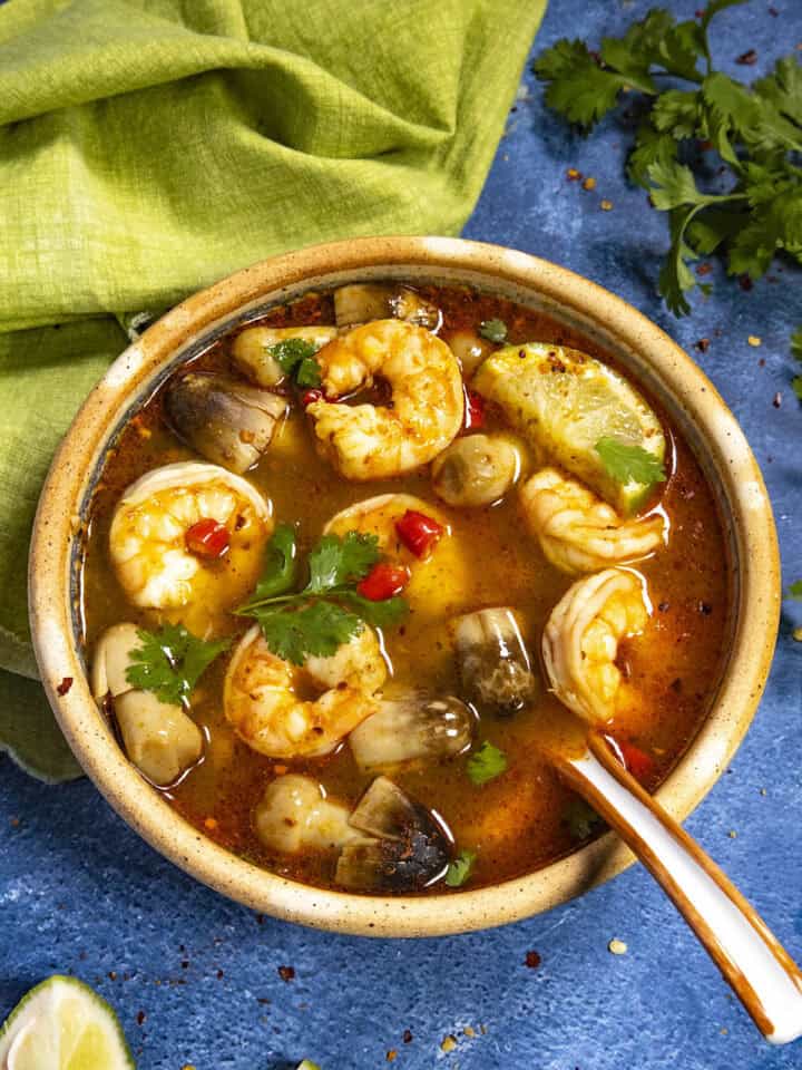 A large bowl of spicy Tom Yum Soup loaded up with shrimp and mushrooms, garnished with spicy red chilies, cilantro, and lime