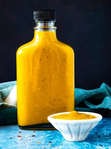 A bottle of spicy, vibrant yellow Bajan Hot Sauce on a table with a bowl of hot sauce beside it