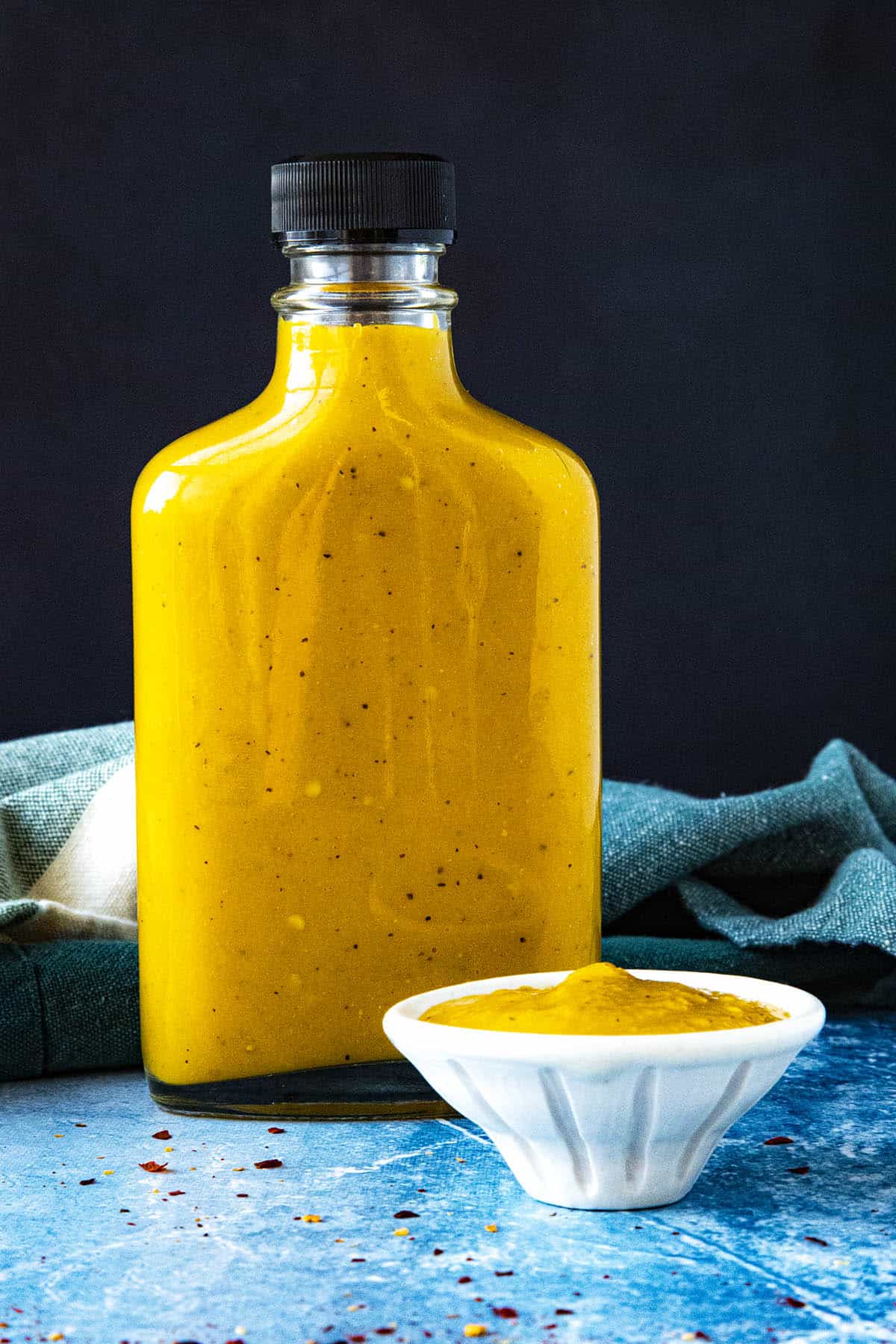 A bottle of spicy, vibrant yellow Bajan Hot Sauce on a table with a bowl of hot sauce beside it