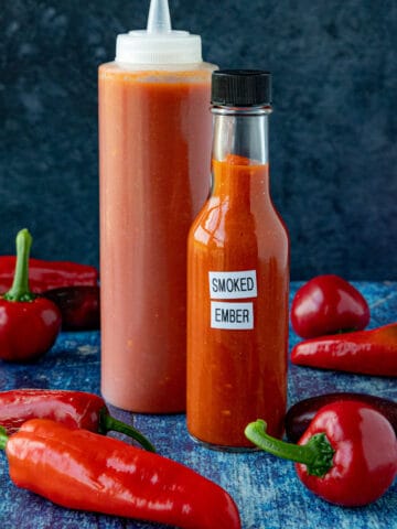 2 bottles of Smoked Chili Hot Sauce (Smoked Ember) on a table with extra chili peppers