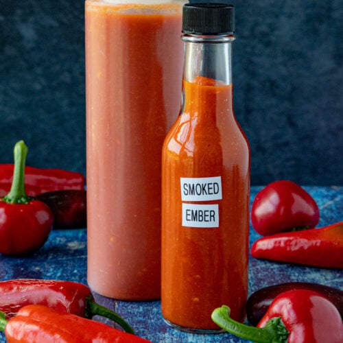 2 bottles of Smoked Chili Hot Sauce (Smoked Ember) on a table with extra chili peppers