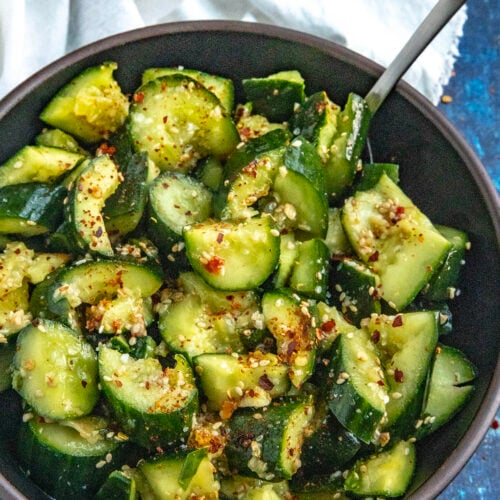 A bowl of Asian Cucumber Salad with a serving spoon, garnished with sesame seeds and chili flakes