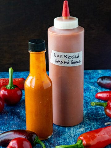 2 bottles of Sun-Kissed Umami Hot Sauce on a table surrounded by freshly picked chili peppers from my garden