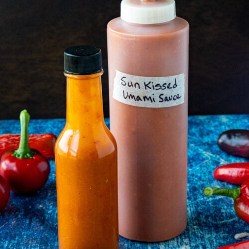 2 bottles of Sun-Kissed Umami Hot Sauce on a table surrounded by freshly picked chili peppers from my garden