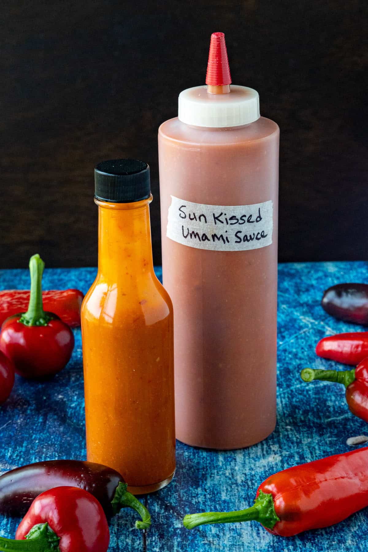 2 bottles of Sun-Kissed Umami Hot Sauce on a table surrounded by freshly picked chili peppers from my garden
