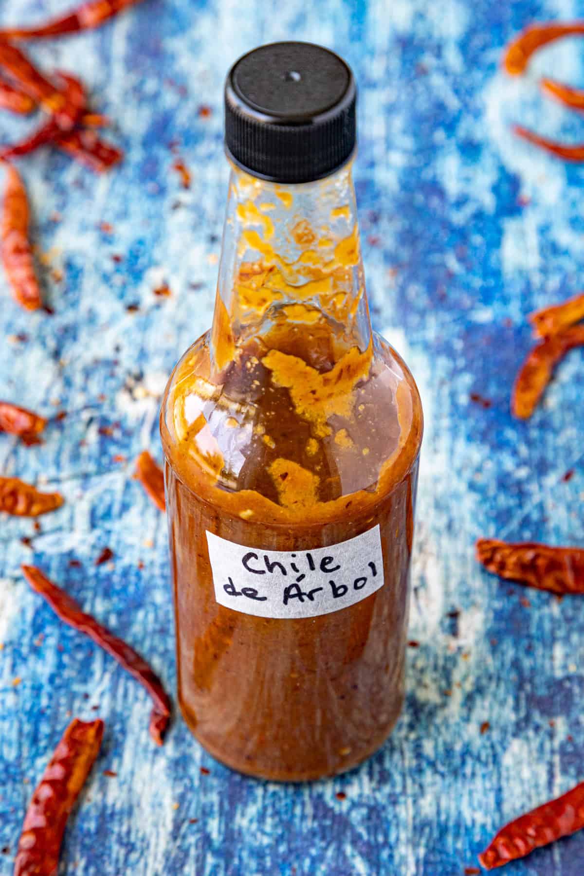 A bottle of fiery Chili de Árbol Hot Sauce surrounded by dried chilies