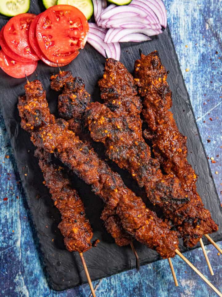 Spicy African Grilled Beef Skewers (Suya) served on a platter with sliced red onion, tomatoes, and cucumbers
