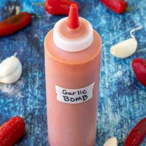 A full bottle of homemade Garlic Bomb Hot Sauce surrounded by fresh garlic and red chilies