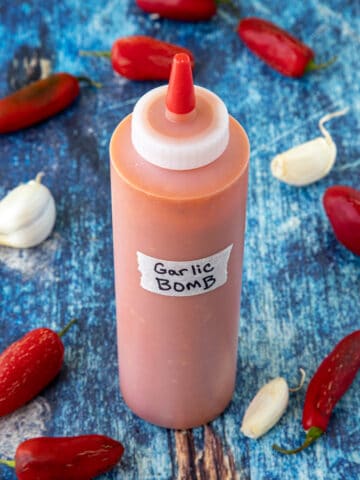 A bottle of homemade Garlic Bomb Hot Sauce surrounded by fresh garlic and red chilies