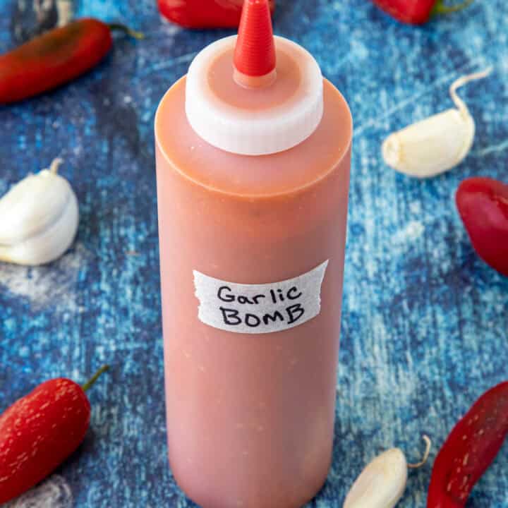 A bottle of homemade Garlic Bomb Hot Sauce surrounded by fresh garlic and red chilies