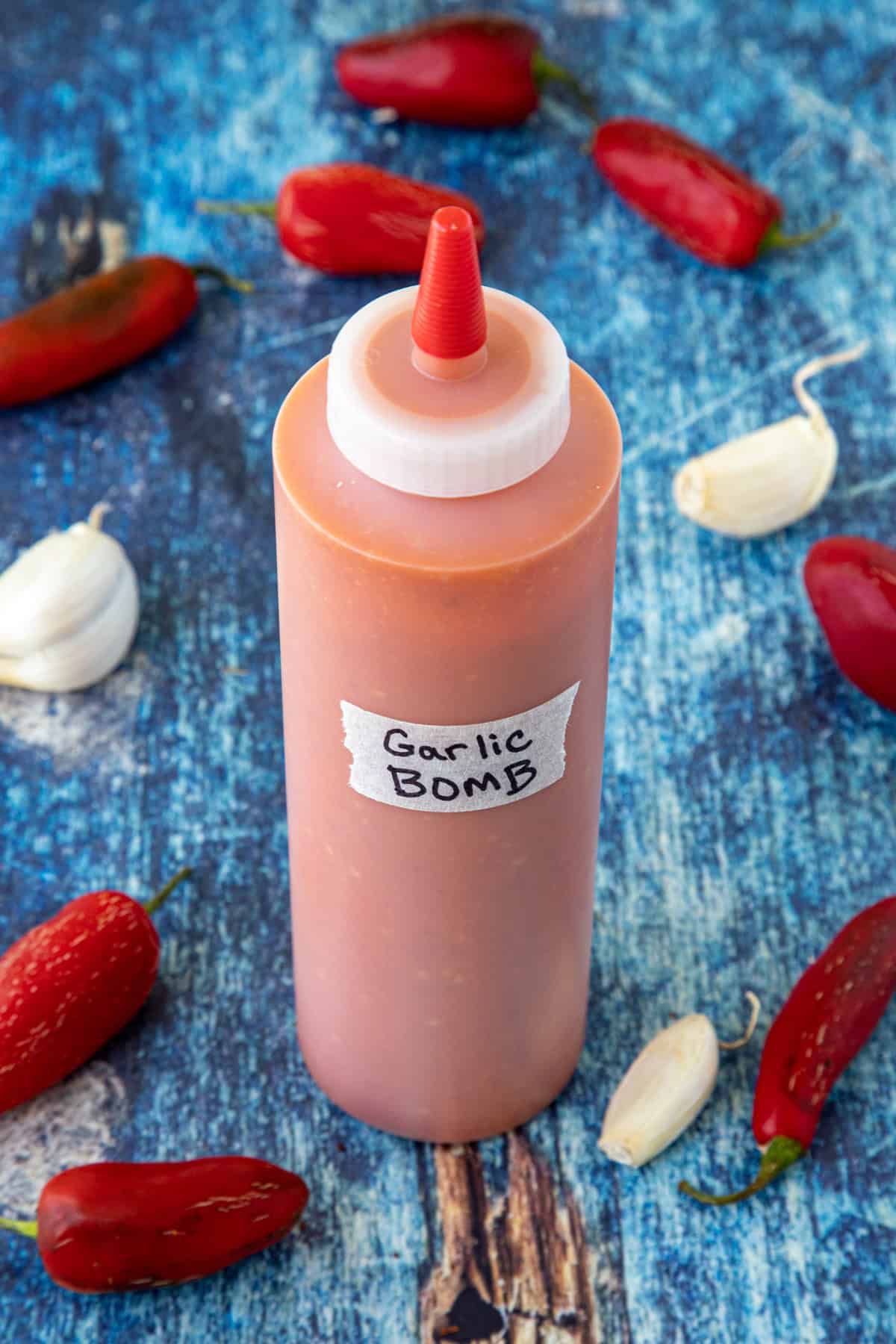 A bottle of homemade Garlic Bomb Hot Sauce surrounded by fresh garlic and red chilies