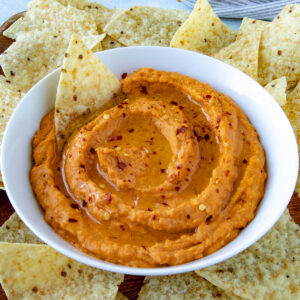 A bowl of creamy white bean dip with harissa topped with olive oil and red pepper flakes, surrounded by tortilla chips on a wooden surface.