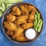 A plate of crispy Buffalo Wings garnished with herbs, served with celery sticks and a small cup of ranch dipping sauce on a blue background.