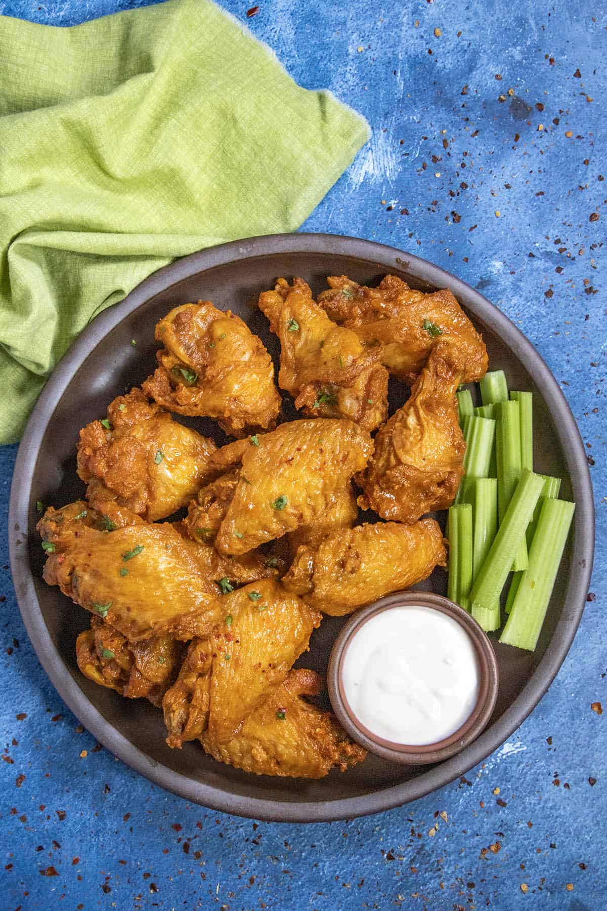 A plate of crispy Buffalo Wings garnished with herbs, served with celery sticks and a small bowl of ranch dipping sauce, sits on a blue textured surface with a green napkin nearby.