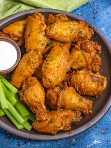 A plate of golden-brown Buffalo Wings garnished with herbs, served with celery sticks and a small bowl of ranch dipping sauce, sits on a dark plate against a blue background.