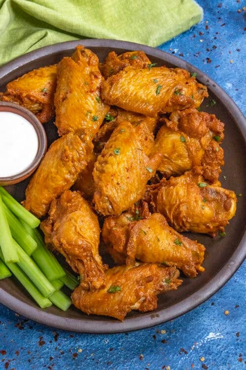 A plate of golden-brown Buffalo Wings garnished with herbs, served with celery sticks and a small bowl of ranch dipping sauce, sits on a dark plate against a blue background.