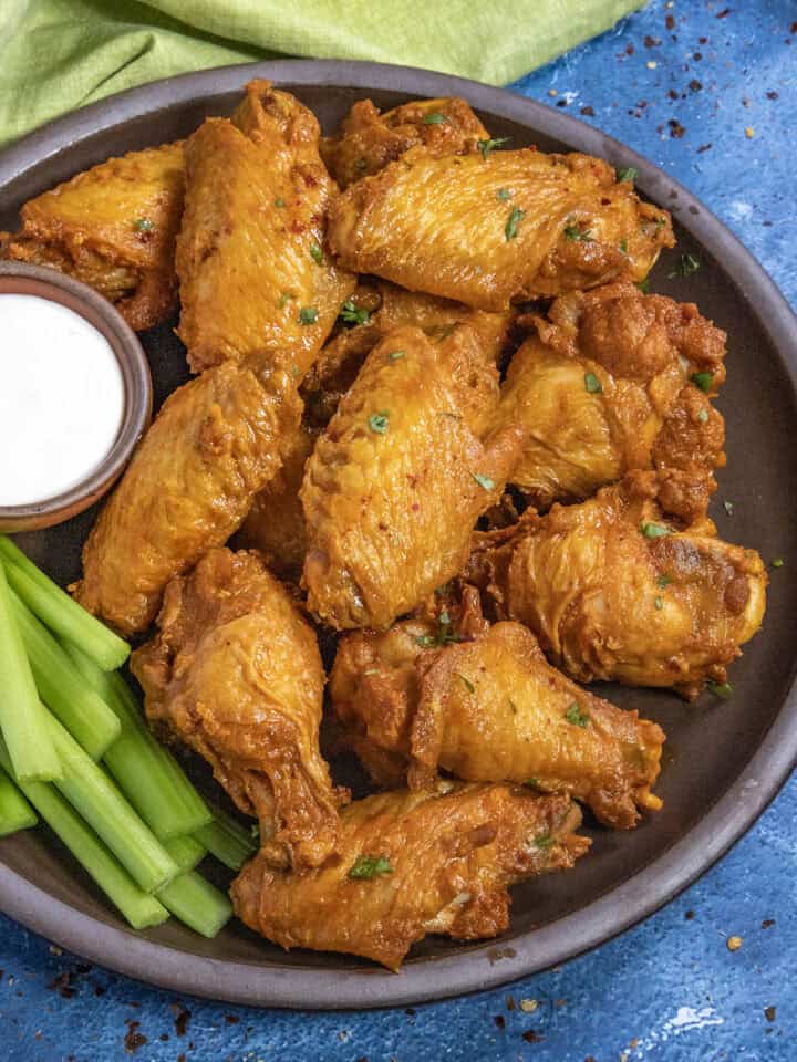 A plate of golden-brown Buffalo Wings garnished with herbs, served with celery sticks and a small bowl of ranch dipping sauce, sits on a dark plate against a blue background.