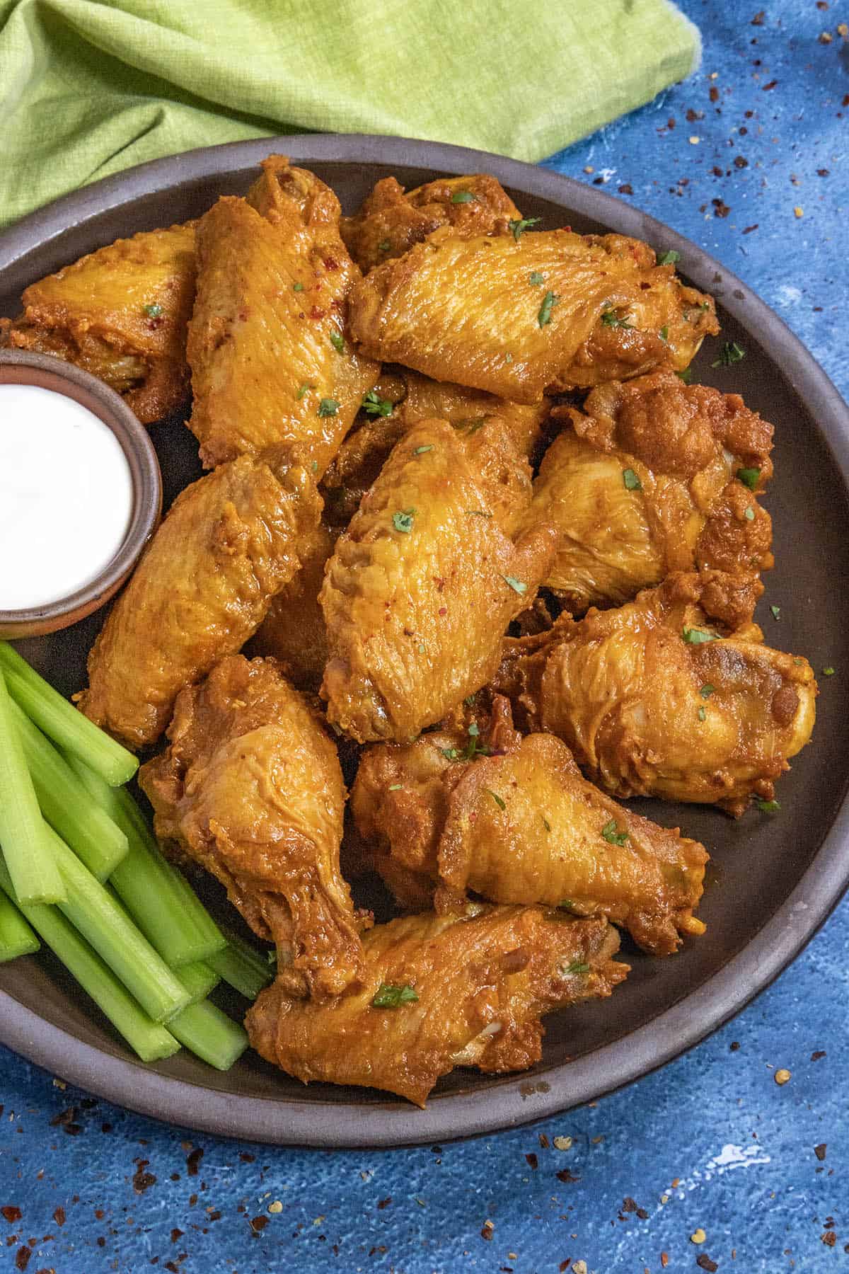 A plate of golden-brown Buffalo Wings garnished with herbs, served with celery sticks and a small bowl of ranch dipping sauce, sits on a dark plate against a blue background.