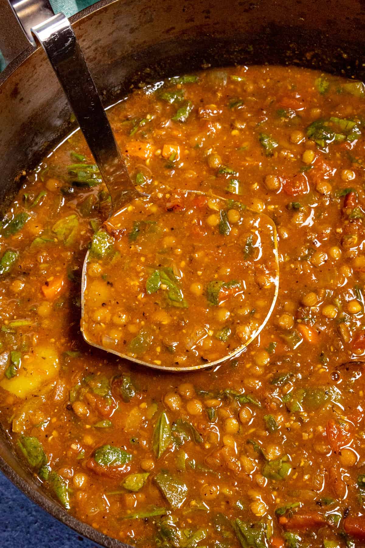 A close-up of a pot filled with thick Lentil Soup, packed with chopped vegetables and greens. A metal ladle lifts a portion of the hearty, tomato-based soup from the pot.