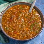 A bowl of hearty Lentil Soup with visible vegetables and herbs, garnished with fresh parsley, sits on a blue surface beside a green cloth. A spoon rests in the flavorful soup.